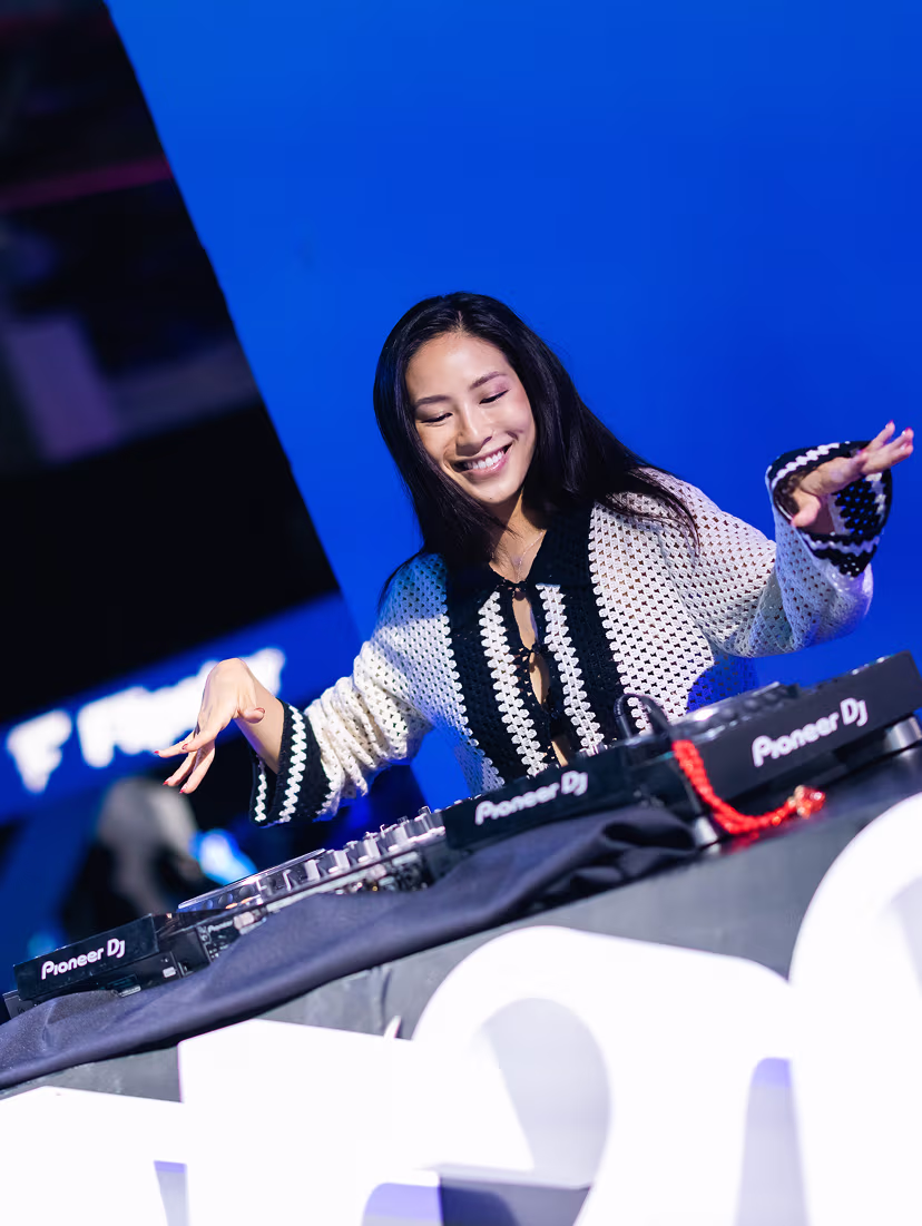 Smiling female DJ performing with Pioneer DJ equipment on a stage with a blue background.